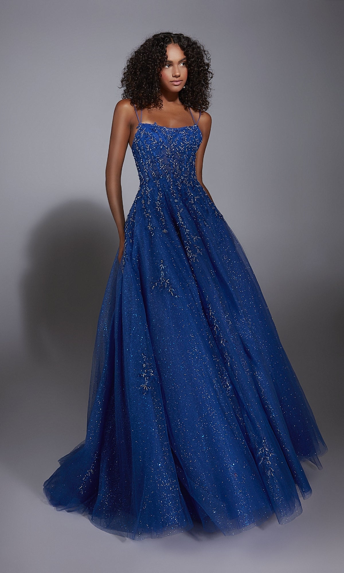 A woman wears the Alyce 61824 Long Prom Dress, a blue floor-length ball gown with a corset bodice, thin straps, and sparkly embellishments, standing against a plain gray background.